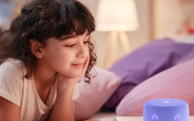 Девочка лежит на кровати и смотрит на умную колонку с экраном